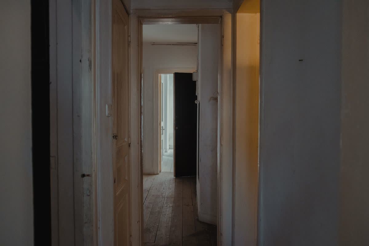 Dim hallway with gray floorboards, a pale door and white switch on the left, and a dark door farther back; no readable text or faces.